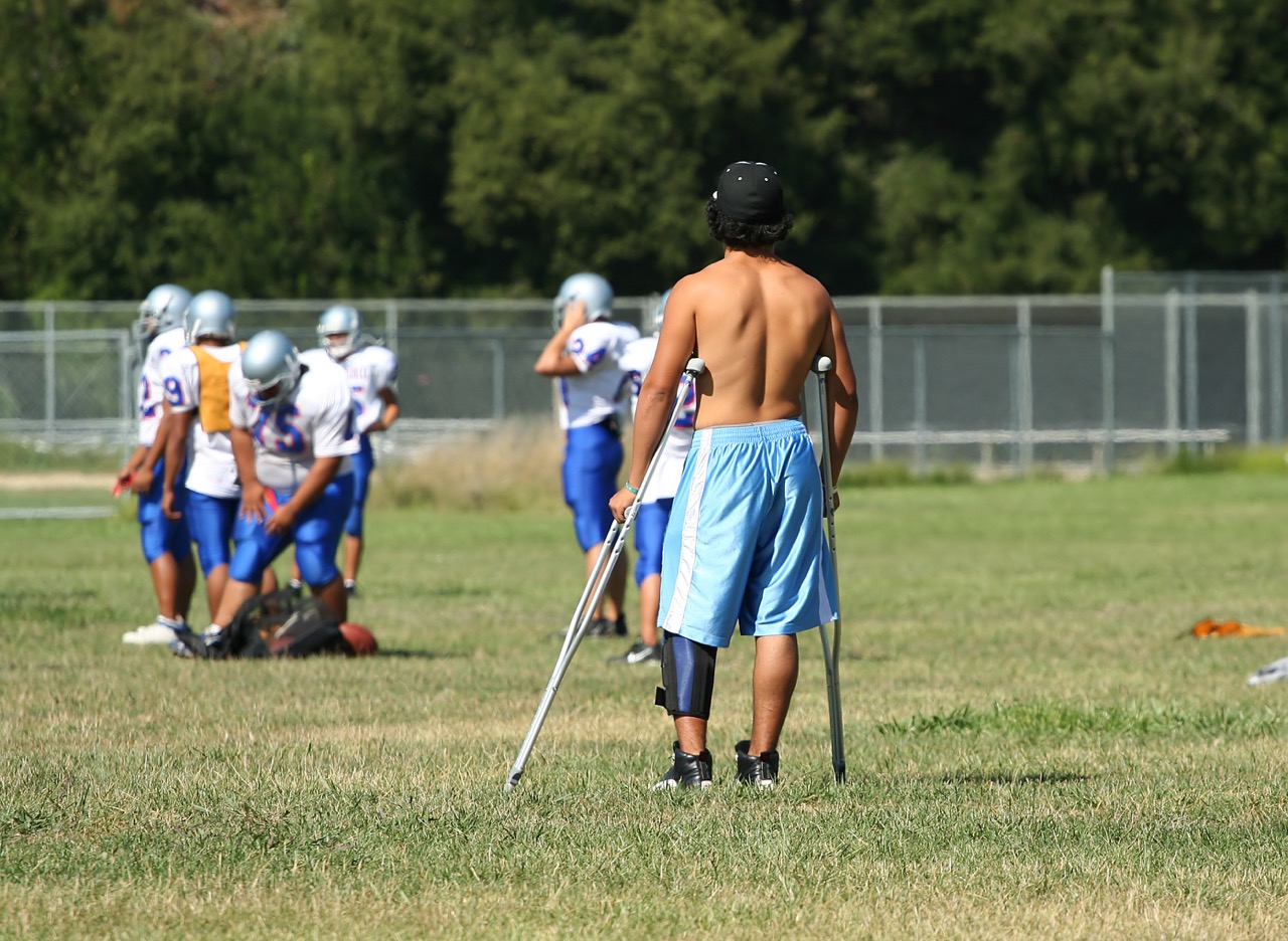 n injured college football player on crutches watches as his team practices without him.